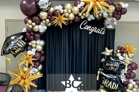 Florida Tech College Graduation Balloon decor and backdrop in Indialantic FL. Maroon and chrome gold organic balloon garland on a black fabric backdrop with grad balloons and gold starburst balloons. Roman style balloon columns and balloon topiary were pl