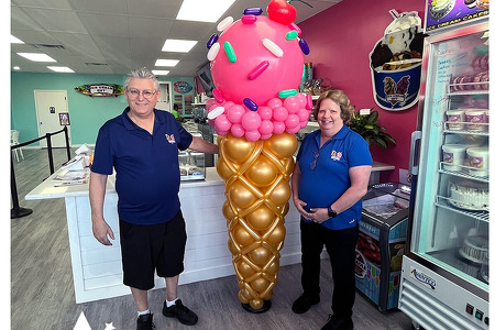 7 foot tall ice cream balloon sculpture with pink ice cream and pastels sprinkles in a gold cone with a cherry on top. This pictures features the owners standing on each side inside their ice cream parlor The Ice Cream Hut on Babcock Road in Palm Bay