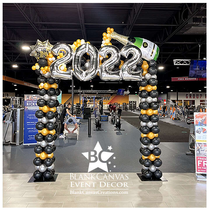 Large Square Balloon Arch with stirpes of gold, black and silver with a large silver 2022 across the top and a huge champagne bottle balloon on top flowing with gold and white bubble champagne and a Happy New Years balloon in the bubbles